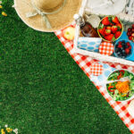 Sommerliches Picknick auf dem Rasen mit offenem Picknickkorb, Obst, Salat und Kirschkuchen