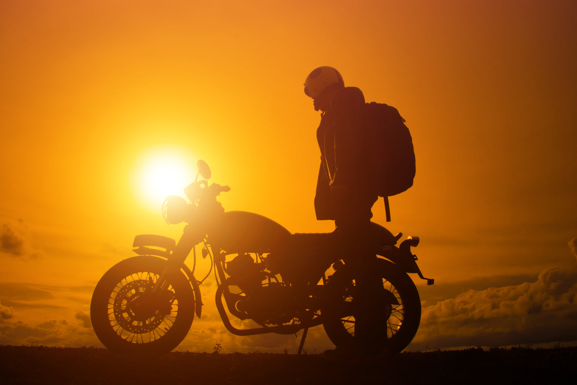 Silhouette eines Bikers mit seinem Motorrad, er trägt einen Schulterrucksack, Sonnenuntergang im Hintergrund.